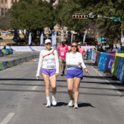 Two women wearing white tops and purple shorts walk side by side on a sunny street during a race event, showcasing their endurance as they navigate colorful barriers and other participants in the background. Austin Marathon Half Marathon & 5K