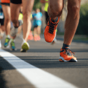 Close-up of runners' legs and feet during a race, with one in bright orange shoes leading the way. The image captures energy and motion, inspiring you to increase mileage and build running distance while avoiding burnout. Austin Marathon Half Marathon & 5K