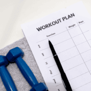 A workout plan sheet with columns for exercises, weight, and reps lies on a gray towel next to two blue dumbbells and a black pen—perfect for those looking to build running distance while tracking progress throughout the week. Austin Marathon Half Marathon & 5K