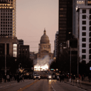 A city street at dusk leads to a domed capitol building, with crowds and cars lining the road. Bright lights and smoke hint at a Race Weekend celebration, making it the perfect scene for an unforgettable Austin Vacation. Austin Marathon Half Marathon & 5K