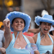 Two women wearing light blue cowboy hats and matching outfits are smiling and celebrating at an outdoor event. One points at the camera as they cheer excitedly during the 24 Weeks countdown to the Austin Marathon, their hats glowing with lights and feather trim. Austin Marathon Half Marathon & 5K