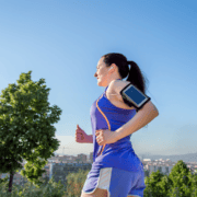 A woman in sportswear jogs outdoors on a sunny day, following her half marathon training plan. She wears earphones and an armband with a phone, with trees and a cityscape behind her under a clear blue sky. Austin Marathon Half Marathon & 5K