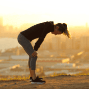 A woman in athletic clothing stands outdoors at sunset, bent over with hands on knees, appearing tired after training. A cityscape and body of water are visible in the blurred background, hinting at the importance of burnout prevention. Austin Marathon Half Marathon & 5K