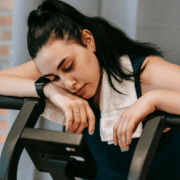 A woman with dark hair and a towel around her neck leans tiredly on a treadmill, looking down with a fatigued expression—a reminder of the importance of burnout prevention during intense training. Austin Marathon Half Marathon & 5K
