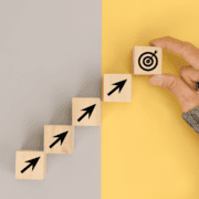 A hand places a wooden block with a target icon at the top of a staircase made of blocks with upward-pointing arrows, symbolizing the 24 weeks countdown to the Austin Marathon, set against a split gray and yellow background. Austin Marathon Half Marathon & 5K