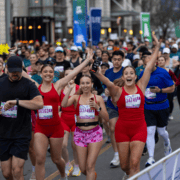 A group of runners participate in the Austin Marathon, with three women in the front smiling and raising their arms in celebration. Many participants are visible behind them, running on a city street and sharing in the exhilarating race experience. Austin Marathon Half Marathon & 5K