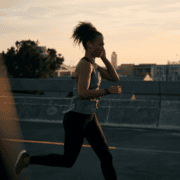 A woman jogs on a city street at sunset, wearing athletic clothes and earphones—embracing February planning for a balanced lifestyle—with buildings and trees visible in the background. Austin Marathon Half Marathon & 5K