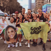 A group of smiling runners pose together after a race, holding medals, a “GOOD GRIEF” flag, and a large cutout of a woman’s face. Tall buildings and other runners are visible in the background, capturing the energy and camaraderie that follow good time management and careful February planning. Austin Marathon Half Marathon & 5K