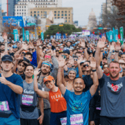 A large group of marathon runners, wearing race bibs and colorful athletic gear, smile, wave, and raise their hands as they gather at the starting line in a city street—perfectly embodying the excitement and disciplined time management that goes into successful February planning. Austin Marathon Half Marathon & 5K