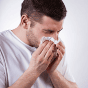 A man in a white shirt is holding a tissue to his nose and appears to be sneezing or blowing his nose, highlighting the importance of running safety tips—especially when you train safely in heat, as allergies and heat can both impact your performance. Austin Marathon Half Marathon & 5K