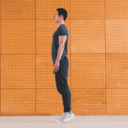 A man stands in profile on his toes with arms at his sides, dressed in a dark t-shirt, joggers, and white sneakers, against a wooden panel wall—the perfect pose to showcase essential workouts that promote both strength and marathon longevity. Austin Marathon Half Marathon & 5K