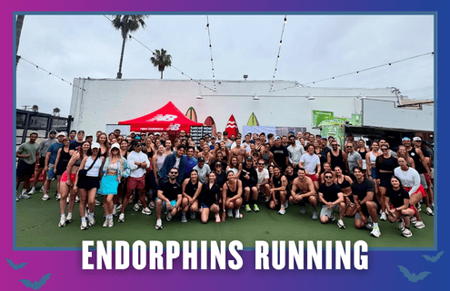 A large group of people pose outdoors in athletic wear under string lights, with surfboards and a New Balance tent in the background. Text at the bottom reads “ENDORPHINS RUNNING.” This spirited Group Run was part of TEAM FX’s KickOff Week celebration. Austin Marathon Half Marathon & 5K