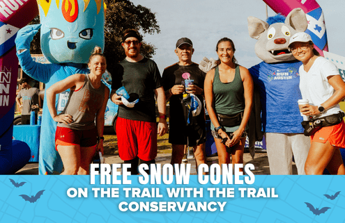 A group of people and two mascots stand smiling outdoors at a fun run event. A blue banner at the bottom reads, “Free snow cones on the trail with the Trail Conservancy,” highlighting this free event for all participants. Austin Marathon Half Marathon & 5K
