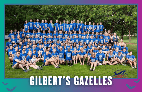 A large group of people wearing matching blue shirts sits and stands together outdoors on grass, smiling at the camera during a Group Run. The text “GILBERT’S GAZELLES” is displayed at the bottom of the image. Austin Marathon Half Marathon & 5K