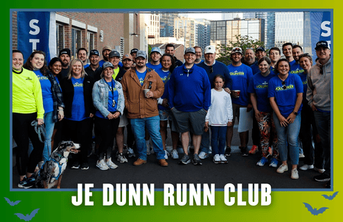 A group of people, some wearing blue “JE Dunn Runn Club” shirts, pose together outdoors near a building in South Austin. A dog is sitting in front. Text at the bottom reads “JE DUNN RUNN CLUB,” highlighting the excitement of kickoff week for their group run. Austin Marathon Half Marathon & 5K