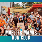 A diverse group of smiling women pose together outdoors, some wearing rainbow accessories and athletic gear, holding a sign that reads “Mueller Mamis Run Club.” Two small dogs are also present, celebrating the kickoff week of their marathon club with this joyful group run. Austin Marathon Half Marathon & 5K