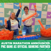 Two people smiling behind a table with PNC Bank swag at the Austin Marathon Health & Fitness Expo. Banners and orange promo items are displayed, with text announcing PNC Bank as the marathon’s official banking partner. Austin Marathon Half Marathon & 5K
