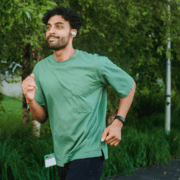 A man wearing a green shirt, black shorts, and wireless earbuds jogs outdoors on a path surrounded by trees and greenery, making the most of his upgraded running gear for an enhanced training experience. Austin Marathon Half Marathon & 5K