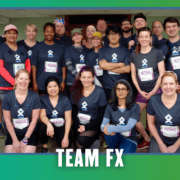 A group of people in matching “TEAM FX” shirts pose indoors, smiling after a Group Run during KickOff Week. They wear race bibs and medals, celebrating their achievement with Goodpain Athletics. The image has a green and blue border with “TEAM FX” written at the bottom. Austin Marathon Half Marathon & 5K