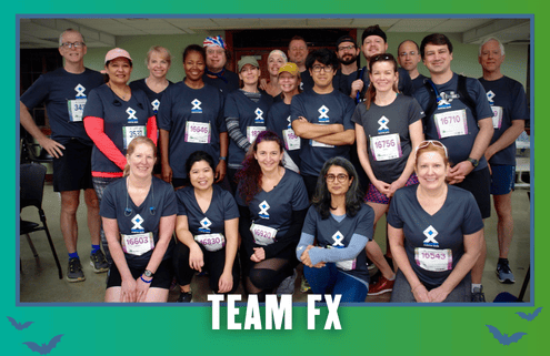A group of people in matching “TEAM FX” shirts pose indoors, smiling after a Group Run during KickOff Week. They wear race bibs and medals, celebrating their achievement with Goodpain Athletics. The image has a green and blue border with “TEAM FX” written at the bottom. Austin Marathon Half Marathon & 5K