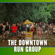 A large group of people poses outdoors at night in running gear in front of a yellow tent and sign, celebrating KickOff Week. Text at the bottom reads, “The Downtown Run Group.” Trees are visible in the background. Austin Marathon Half Marathon & 5K