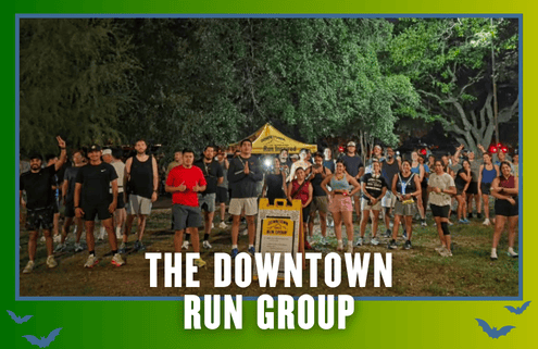 A large group of people poses outdoors at night in running gear in front of a yellow tent and sign, celebrating KickOff Week. Text at the bottom reads, “The Downtown Run Group.” Trees are visible in the background. Austin Marathon Half Marathon & 5K
