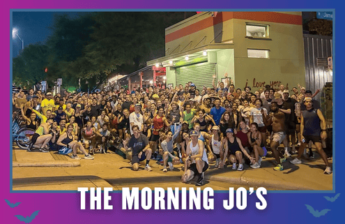 A large group of people pose together outside a building at night. Many are in athletic wear, some crouch in front, and there’s a mural on the wall. Text at the bottom reads “THE MORNING JO’S.” It looks like a celebratory moment from Redemption Run Crew’s KickOff Week Group Run. Austin Marathon Half Marathon & 5K