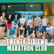 Thirteen runners pose together in athletic gear outside a building, ready for a group run. “Twenty-Six Two Marathon Club” is written at the bottom. The photo, possibly taken during KickOff Week with Mueller Run Club, has a green and blue border with small bat icons in the corners. Austin Marathon Half Marathon & 5K