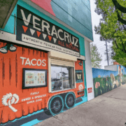 A vibrant mural of an orange truck decorates the exterior of Veracruz All Natural, a taco stand serving authentic Mexican food, smoothies, and juices—a favorite spot for post-run tacos among East Austin running clubs. A sidewalk and mural-covered wall are visible beside the building. Austin Marathon Half Marathon & 5K