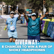 Two runners in blue shirts cheer as they approach the finish line of a race, capturing a moment perfect for a Finisher Pix photo package. A banner at the bottom reads, “GIVEAWAY: 8 CHANCES TO WIN A PAIR OF SHOKZ HEADPHONES.”. Austin Marathon Half Marathon & 5K