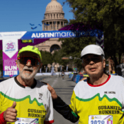 Two male runners in Gusanito Runners jerseys stand together at the Austin Marathon finish line in front of the Texas State Capitol, representing their local running crew. Austin Marathon Half Marathon & 5K