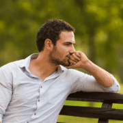 A man with short dark hair sits on a wooden bench outdoors, resting his chin on his hand and looking thoughtful—perhaps reflecting on how to handle a missed run. He wears a light-colored shirt; blurred green foliage fills the background. Austin Marathon Half Marathon & 5K